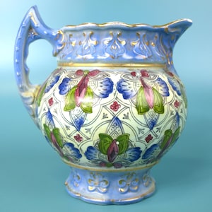 May include: A ceramic jug featuring a light blue base colour with a scalloped rim and a curved handle. The body is decorated with a repeating lattice pattern in white, accented by floral motifs in shades of blue, green, and magenta, with gold trim.