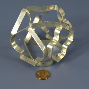 May include: A silver metal cookie cutter shaped like a cube. The cutter is made of multiple pieces of metal that are joined together. The cutter is about the size of a penny.