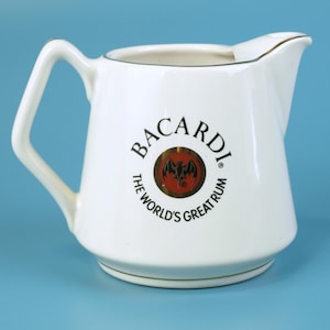 Può includere: Brocca in ceramica bianca con manico e beccuccio. La brocca presenta il logo Bacardi in un design circolare con un pipistrello e la scritta "THE WORLD'S GREAT RUM". Il bordo e la base hanno un bordo dorato. La brocca è su sfondo blu.