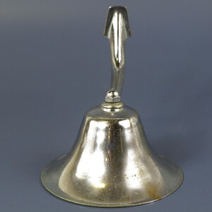 May include: A silver-colored bell with a curved handle and a flared base. The handle extends upwards and curves gracefully to meet the bell's body. The bell has a smooth, reflective surface.