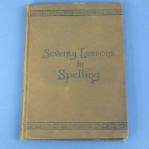 May include: A vintage book titled "Seventy Lessons in Spelling" with a tan cover. The title is in dark blue script. Decorative border lines are at the top and bottom of the cover. The book appears to be old and well-used.