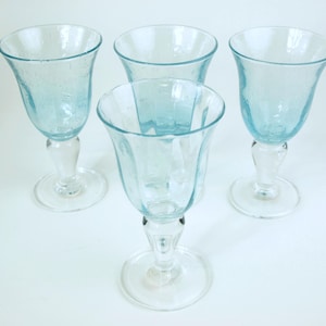 May include: Four light blue stemmed wine glasses with a textured surface. The glasses have a clear, rounded base and a flared bowl. The stem is clear and has a decorative shape. The glasses are arranged on a white surface.