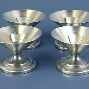 May include: Four silver-colored, stemmed bowls with a wide, shallow bowl and a small, rectangular insert. The bowls have a pedestal base and are arranged on a blue surface. The bowls appear to be made of metal.