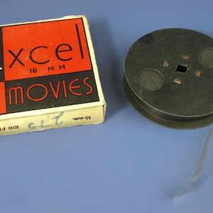 Könnte beinhalten: Vintage Excel 16 MM MOVIES Filmrolle und -box. Die Box ist weiß mit einem roten und schwarzen Design und dem Text "Excel MOVIES". Die Filmrolle ist dunkelgrün und hat einen Filmstreifen, der sich davon erstreckt. Die Box liest "100 FEET".