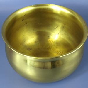 Puede incluir: Un cuenco dorado brillante con forma redondeada. El cuenco tiene una abertura ancha y una superficie lisa y pulida. El interior muestra algunas decoloraciones menores. El cuenco probablemente está hecho de latón o un metal similar.