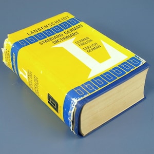 May include: A yellow and blue Langenscheidt Standard German Dictionary. The book has a yellow cover with blue accents and the text "STANDARD GERMAN DICTIONARY" and "GERMAN ENGLISH ENGLISH GERMAN". The book is closed and lying on a blue surface.
