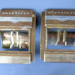 以下が含まれることがあります： ブラウン仕上げとシルバー調のアクセントが施された、ヴィンテージの金属製シボレーシートベルトバックル2点。各バックルの上部にはシボレーのブランド名とロゴが刻印されており、下部には長方形のリリースボタンが付いています。