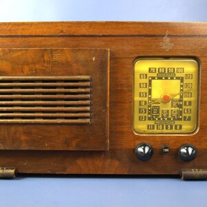 Puede incluir: Una radio vintage de madera con un dial dorado y botones negros. El dial tiene una escala en megaciclos y kilociclos. La radio tiene una rejilla de altavoz en el lado izquierdo.