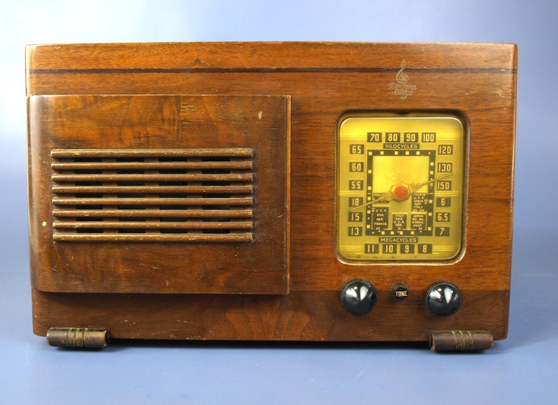 Puede incluir: Una radio vintage de madera con un dial dorado y botones negros. El dial tiene una escala en megaciclos y kilociclos. La radio tiene una rejilla de altavoz en el lado izquierdo.