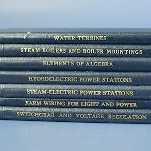 Puede incluir: Una pila de siete libros de tapa dura azul oscuro con letras doradas. Los títulos incluyen "Water Turbines", "Steam Boilers" y "Elements of Algebra". Los libros probablemente están relacionados con la ingeniería o temas técnicos.