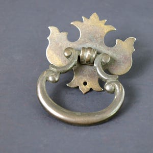 May include: Antique brass drawer pull with a decorative backplate and a curved handle. The backplate has a floral design with a central hole for mounting. The handle is a semi-circular shape. The hardware is tarnished, giving it a vintage look.