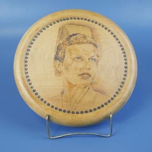 May include: A round wooden plaque featuring a portrait of a woman in a hat, etched into the wood. The portrait is surrounded by a dotted border. The plaque is displayed on a metal stand against a blue background.