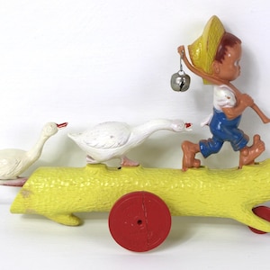 May include: A vintage toy featuring a yellow log-shaped cart with red wheels, carrying two white geese and a boy figure. The boy is wearing blue overalls and a straw hat, carrying a bell.