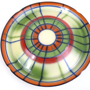 Może przedstawiać: Kolorowy ceramiczny talerz do serwowania o koncentrycznym wzorze. Talerz posiada centralne zagłębienie i szeroki rant. Schemat kolorów obejmuje pomarańczowy, zielony, czerwony, niebieski i kremowy, z wzorem kraty.