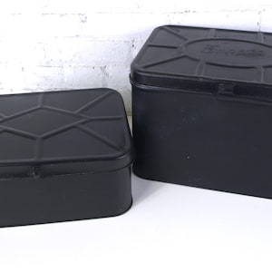May include: Two black rectangular metal storage tins with lids. The larger tin has a raised oval design on the lid with the word "Brigade" embossed. The smaller tin has a diamond pattern on the lid.