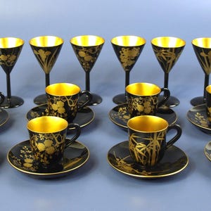 May include: A set of black and gold vintage glassware, including stemmed glasses, teacups with saucers, and a serving dish. The interior of the glasses and cups are gold. Floral and bamboo designs adorn the black surfaces.