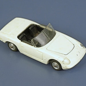 May include: A white toy convertible car with black interior and a windshield. The miniature vehicle has visible wear and tear, including paint chips. The car has small wheels and is set against a blue background.