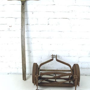 May include: An old push lawn mower with a wooden handle and a rusty metal cutting mechanism. The handle is tall and has a horizontal grip. The mower has two large wheels and a series of blades.
