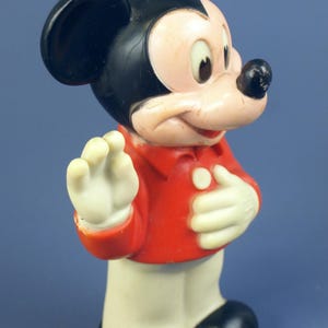 May include: Vintage Mickey Mouse figurine. The figure is wearing a red jacket, white gloves, and black shoes. The classic cartoon character has a black head and ears, a pink face, and a black nose. The figurine is standing and waving.