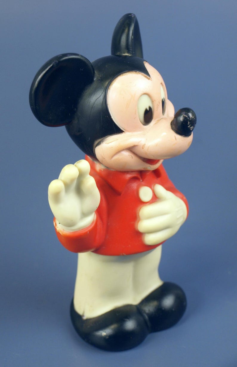 May include: Vintage Mickey Mouse figurine. The figure is wearing a red jacket, white gloves, and black shoes. The classic cartoon character has a black head and ears, a pink face, and a black nose. The figurine is standing and waving.
