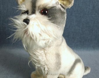 Può includere: Un cane di peluche bianco e grigio con occhi neri e una barba bianca e soffice. Il cane è seduto con le zampe ripiegate sotto il corpo.