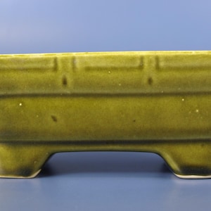 Könnte beinhalten: Ein rechteckiger, olivgrüner Keramik-Pflanztopf mit glänzendem Finish. Der Pflanztopf hat ein abgestuftes Design mit drei kleinen Füßen. Der obere Rand weist eine Reihe von vertieften rechteckigen Details auf. Der Hintergrund ist einfarbig blau.