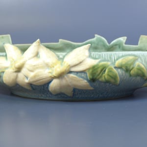 Pode incluir: Uma tigela decorativa de cerâmica azul clara com um design floral. A tigela apresenta flores creme e folhas verdes. A tigela tem alças e mede aproximadamente 30 cm de comprimento.