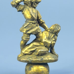 Könnte beinhalten: Kleine, antike Messingfigur, die eine Figur in Rüstung darstellt, die eine andere kniende Figur schlägt. Die Skulptur ist auf einem runden Sockel auf einer Kugel montiert. Die Gesamtfarbe ist ein getrübtes Gold.