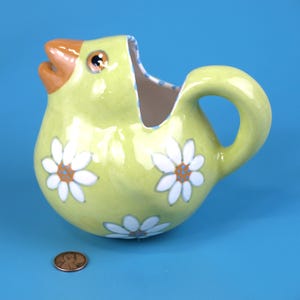 Puede incluir: Una jarra de cer&aacute;mica con forma de pollito amarillo con pico y ojo naranja. Flores de margarita blancas con centros naranjas decoran la jarra. El asa es curva y el interior tiene un estampado de lunares azules y blancos.