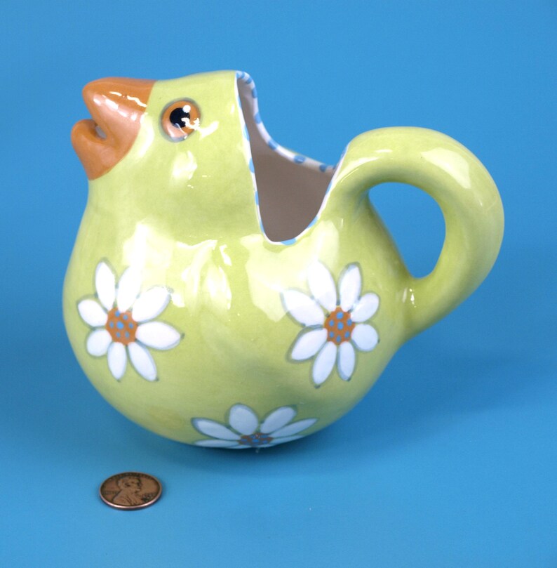 Puede incluir: Una jarra de cer&aacute;mica con forma de pollito amarillo con pico y ojo naranja. Flores de margarita blancas con centros naranjas decoran la jarra. El asa es curva y el interior tiene un estampado de lunares azules y blancos.