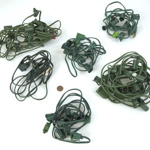 Puede incluir: Varios juegos enredados de luces navideñas verdes con varios estilos de enchufe están esparcidos sobre una superficie blanca. Algunos juegos están enrollados firmemente, mientras que otros están más sueltos. También se ve una moneda.