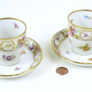 May include: Two white teacups and saucers with a floral pattern in shades of pink, purple, yellow, and green. The cups have gold trim around the rim and a gold band around the middle. The saucers have a gold trim around the edge.
