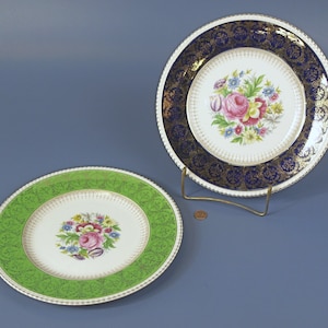 May include: Two decorative plates with floral designs. One plate has a green border with a white center, and the other has a navy blue border with a white center. Both plates feature a central floral arrangement with pink roses and other colorful flowers.