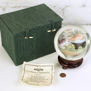May include: A glass paperweight with a hand-painted scene of a cat and a bear in a forest setting. The paperweight is displayed on a wooden base and comes with a certificate of authenticity. The certificate reads "This hand-crafted glass ball is an authentic work, hand-painted as part of the "Priceless Collection." # 4381"