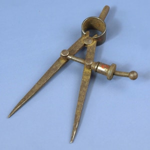 May include: A vintage brass compass with pointed tips and a screw adjustment. The compass has a circular ring at the top and a screw mechanism for adjusting the width. The metal has a worn, aged appearance.