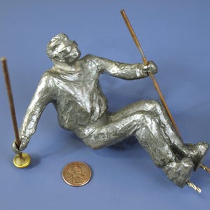 May include: A silver-colored metal figurine of a skier in a seated position, holding a ski pole in each hand. The figurine is mounted on a wooden base with two metal poles.