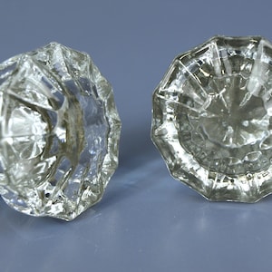 May include: Two clear glass door knobs featuring a multi-faceted, starburst-like design with twelve distinct edges. Each knob has a circular, transparent glass face and a metallic base component visible on the side of one of the knobs.