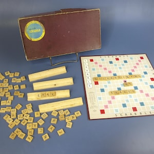 May include: Vintage Scrabble game set with a maroon box, game board, wooden letter tiles, and tile racks. The game board has a grid pattern with colored squares. The word "SILVERLINING BY TRAVIS" is spelled out on the board.