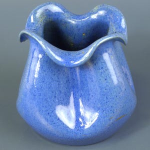 May include: A blue ceramic vase with a ruffled rim. The vase has a speckled texture and a glossy finish. The opening has a unique, wavy shape. The vase is a medium size and has a rounded body.