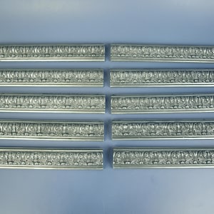 Puede incluir: Diez molduras decorativas plateadas con diseños florales y de volutas ornamentadas. Cada pieza rectangular mide unos 30 cm de largo, dispuestas sobre una superficie azul claro. Para proyectos de interiorismo.