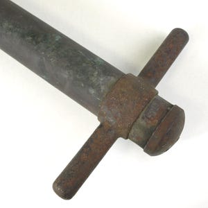 May include: A weathered metal tool featuring a long, cylindrical shaft with a dark, oxidized patina. At one end, a T-shaped handle is attached, showing significant rust and surface corrosion, with a rounded cap securing the assembly in place.
