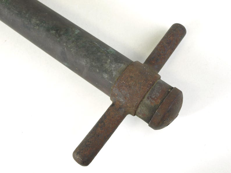 May include: A weathered metal tool featuring a long, cylindrical shaft with a dark, oxidized patina. At one end, a T-shaped handle is attached, showing significant rust and surface corrosion, with a rounded cap securing the assembly in place.