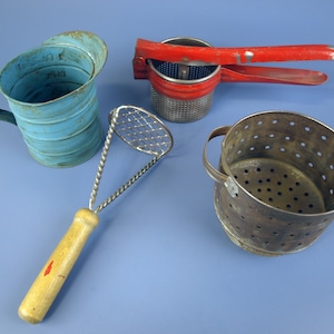Puede incluir: Colección de utensilios de cocina antiguos, que incluye una taza medidora azul, un pasapurés rojo y plateado, un machacador de patatas y un colador de metal. Las herramientas están dispuestas sobre una superficie azul.