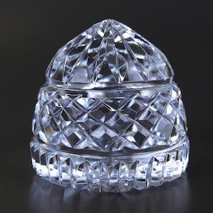 May include: A clear, faceted crystal jar with a domed lid. The jar's body and lid feature a diamond-cut pattern, reflecting light. The jar is likely used for storing small items and has a classic, elegant design.
