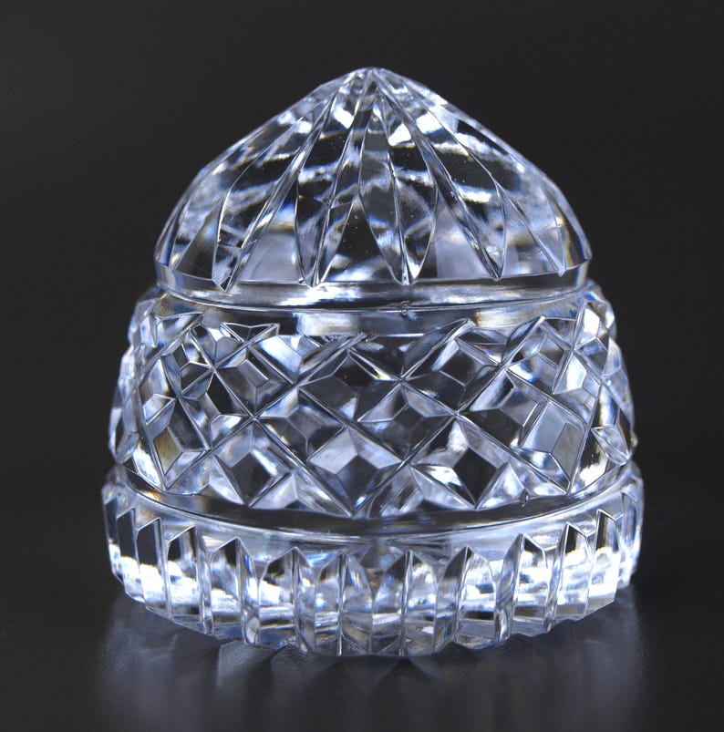 May include: A clear, faceted crystal jar with a domed lid. The jar's body and lid feature a diamond-cut pattern, reflecting light. The jar is likely used for storing small items and has a classic, elegant design.