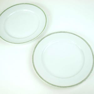 Op de afbeelding: Twee witte keramische borden met een decoratieve lichtgroene rand. Eén bord is ovaal en de andere is rond. De borden staan tegen een witte achtergrond.