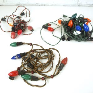 Puede incluir: Tres juegos de luces navideñas vintage. Un juego tiene bombillas rojas y blancas con un cable rojo y negro. Otro juego tiene cable verde con bombillas rojas, naranjas y blancas. El último juego tiene cable marrón con bombillas rojas, verdes y azules.