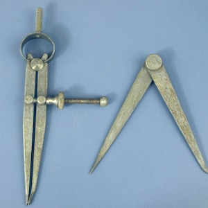 May include: Two vintage metal drafting tools. On the left, a spring-joint divider with a knurled adjustment screw and nut. On the right, a simple friction-joint divider with two pointed legs. Both tools feature a weathered, matte grey finish.