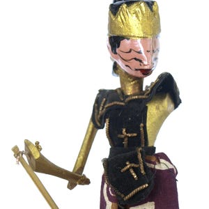 May include: A wooden puppet with a gold crown, black and gold armor, and a brown and white patterned skirt. The puppet is holding a wooden sword and is posed in a fighting stance.