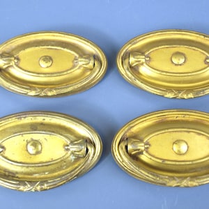 May include: Four oval-shaped brass drawer pulls with a metallic gold finish. Each pull features a central circular mounting point and a hinged drop handle attached to a decorative backplate with a raised border and subtle etched detailing.